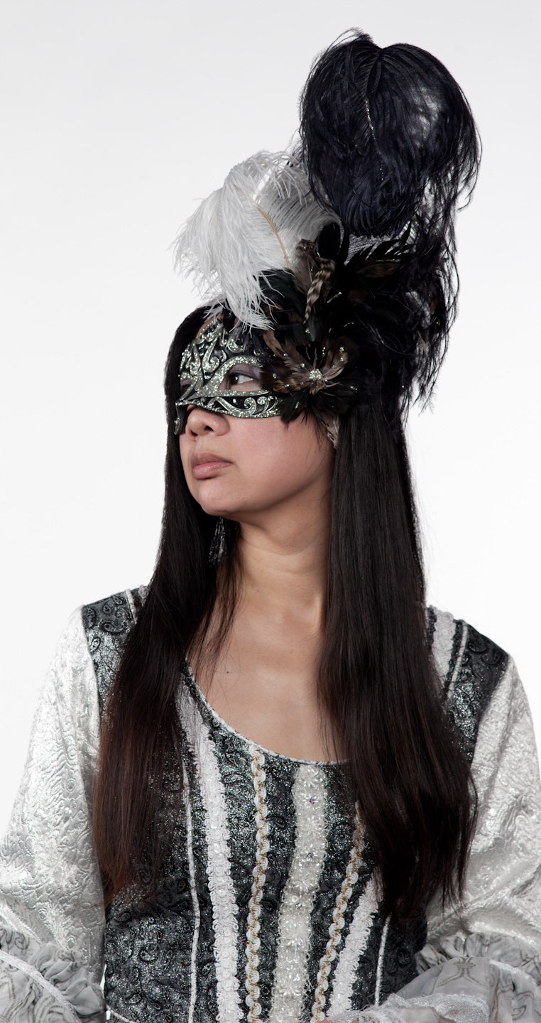 A self portrait of Christina Siu dressed in a black and white Renaissance masquerade costume, complete with a half mask and a plume of upright feathers. She is looking left.